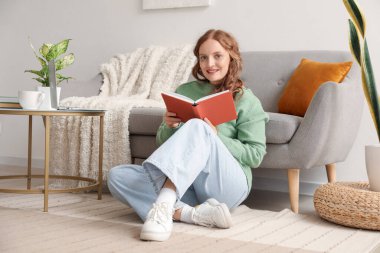 Güzel kızıl saçlı kadın evde kitap okuyor.