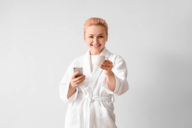 Hafif arka planda bir fincan kahve ve cep telefonu olan bornozlu genç bir kadın.