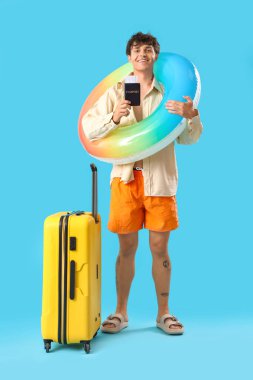 Pasaportu, yüzme yüzüğü ve mavi arka planda bavulu olan genç adam.