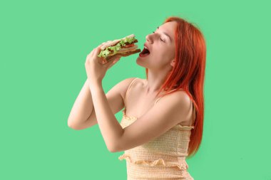 Genç kızıl saçlı kadın yeşil arka planda lezzetli sandviçler yiyor.