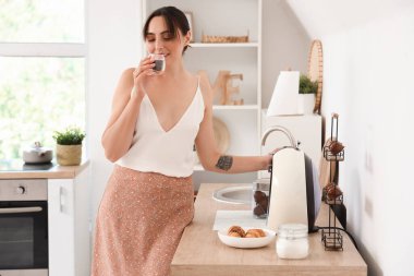 Genç ve güzel bir kadın mutfaktaki kahve makinesinin yanında sıcak espresso içiyor.