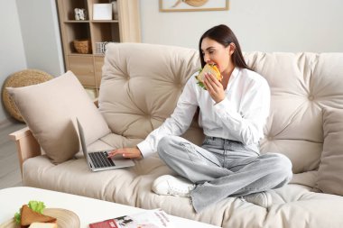 Evde dizüstü bilgisayar kullanan lezzetli sandviçli genç bir kadın.