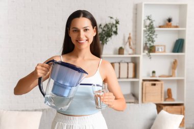 Oturma odasında su filtresi ve cam sürahi olan güzel genç bir kadın.