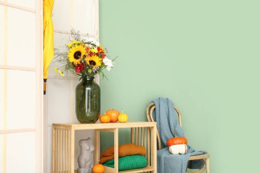 Yeşil duvarın yanında bir buket sonbahar çiçeği, balkabağı ve dekor olan raf ünitesi.