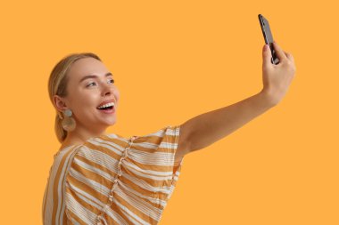 Mutlu genç bir kadın portakal rengi arka planda cep telefonuyla selfie çekiyor.