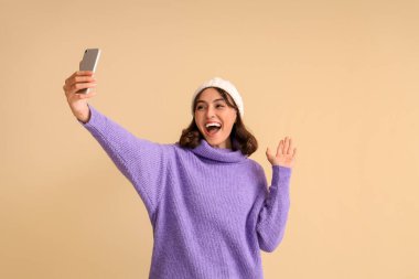 Sıcak kazaklı genç bir kadın bej arka planda selfie çekiyor.