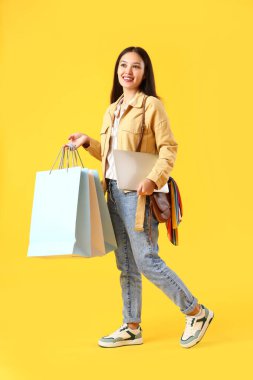 Sarı arka planda alışveriş torbaları ve dizüstü bilgisayarı olan genç Asyalı kadın.