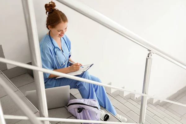Female medical student with clipboard sitting on stairs at university