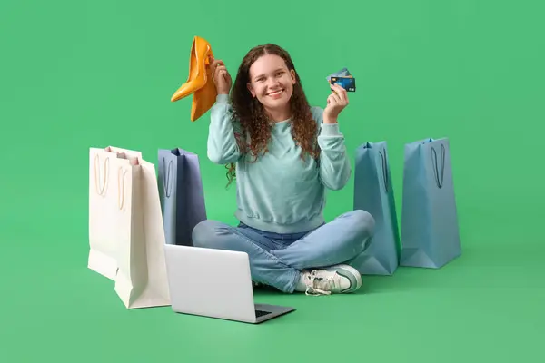 Young woman with shopping bags, laptop, credit cards and new heels sitting on green background