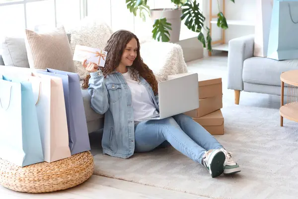 Young woman with laptop, bags and gift card shopping online at home