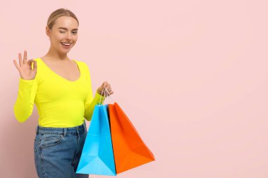 Alışveriş torbalı mutlu genç kadın pembe arka planda iyi niyet gösterisi gösteriyor.