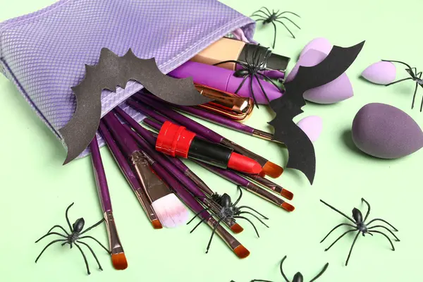 Makeup bag with lipstick, different decorative cosmetics and Halloween decor on green background