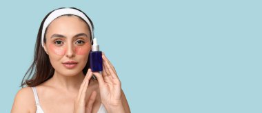 Göz altı bantları ve açık mavi arka planda bir şişe kozmetik yağı olan güzel genç bir kadın.