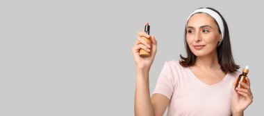 Gri arka planda farklı kozmetik ürünleri tutan güzel genç bir kadın.