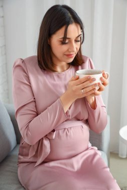 Evdeki kanepede bir fincan çayla oturan hamile bir kadın.