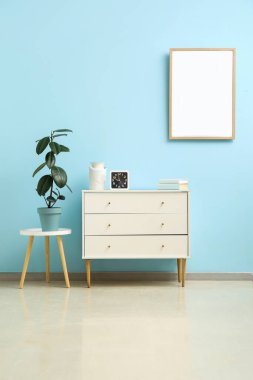 Süslü beyaz çekmeceler, saat ve mavi duvarın yanındaki taburede saksı.
