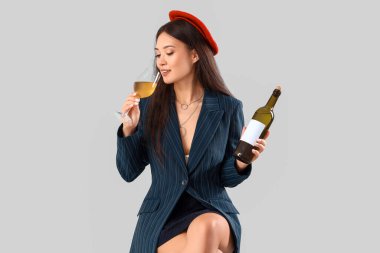 Cam ve bir şişe şarapla gri arka planda oturan güzel Asyalı kadın.