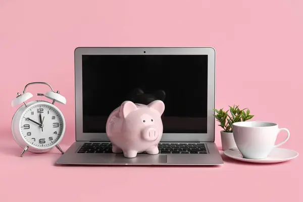 Piggy bank with laptop, cup and alarm clock on pink background