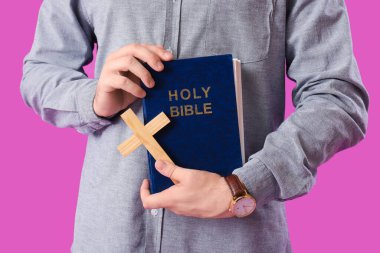 İncil 'i ve ahşap haçı olan genç adam mor arka planda, yakın plan.