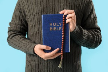 Mavi arka planda İncil ve tespih taşıyan genç adam, yakın plan.