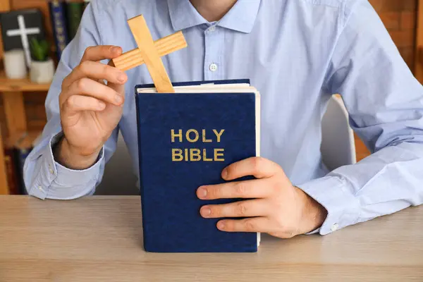Young man with wooden cross and Bible sitting at wooden table, closeup