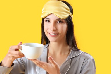 Sarı arka planda kahve fincanı olan pijamalı genç bayan, yakın plan.
