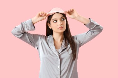 Pembe arka planda uyku maskesi olan pijamalı genç bir kadın.