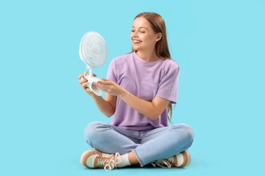 Mavi arka planda oturan elektrikli fanı olan genç bir kadın.