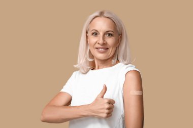 Kolunda yara bandı olan olgun bir kadın bej arka planda baş parmağını kaldırmış. Aşılama kavramı