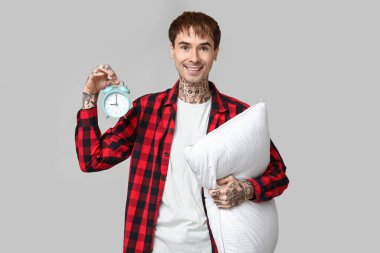 Gri arka planda yastıklı ve çalar saati olan pijamalı dövmeli genç bir adam.