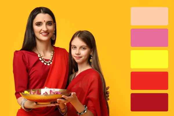 美しいインドの女性と彼女の娘は黄色の背景にキャンドルと花でプレートを保持しています 異なった色のパターン — ストック写真