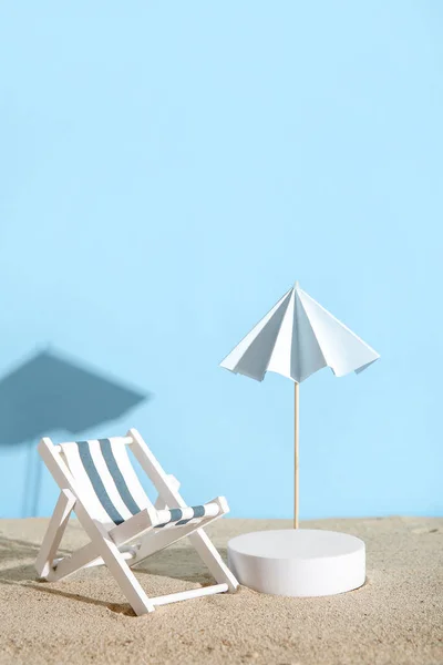 Creative composition with decorative plaster podium, deckchair and umbrella on sand against blue background