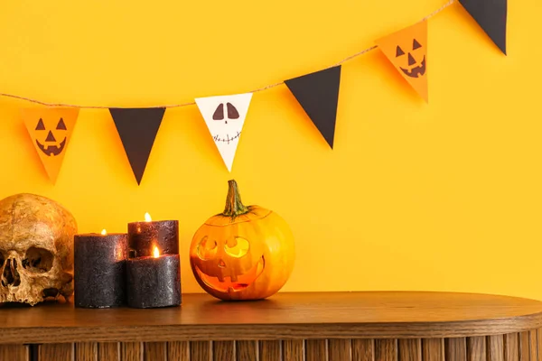 Chest of drawers with burning candles, pumpkin, skull for Halloween celebration and flags hanging on orange wall in room, closeup