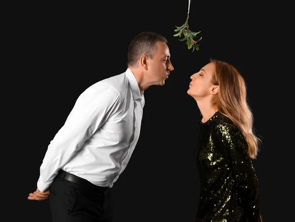 Mature couple kissing under mistletoe branch on dark background