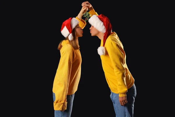Junges Paar Nikolausmützen Küsst Sich Unter Mistelzweig Auf Dunklem Hintergrund — Stockfoto