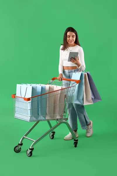 Young woman with tablet computer, shopping cart and bags on green background