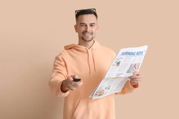 Happy young man with newspaper watching TV on beige background