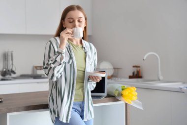 Masanın yanında modern dizüstü bilgisayarı ve sarı lale çiçekleri olan kahve içen güzel bir kadın.