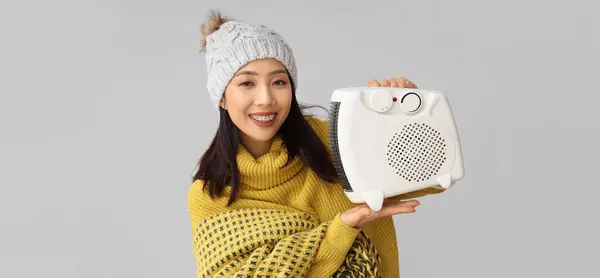 灰色の背景に電気ファンヒーターを保持する若いアジアの女性 ヒーティングシーズン — ストック写真
