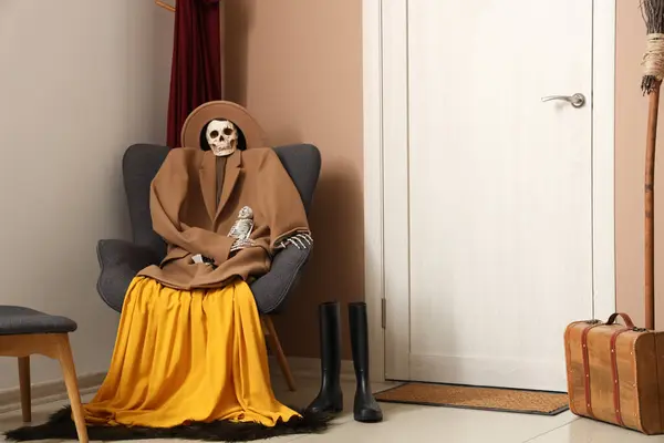 Scary Halloween skeleton in autumn clothes sitting on armchair in living room