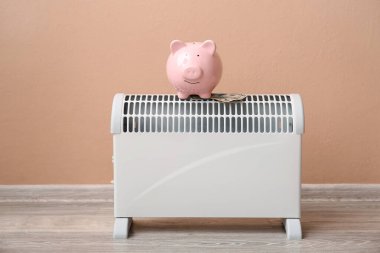 Domuz kumbarası ve evdeki bej duvarın yanındaki elektrikli konteyner ısıtıcısına para. Isıtma kaydetme kavramı
