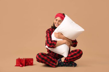 Yastık ve hediye kutularıyla kahverengi arka planda oturan pijamalı güzel bir genç kadın.