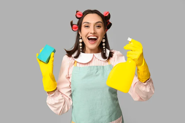 Portrait of happy young housewife in apron with sponge and detergent on grey background