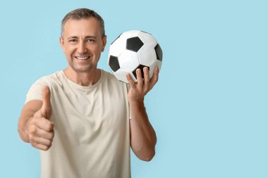 Futbol topuyla yakışıklı, olgun bir adam mavi arka planda baş parmak hareketi yapıyor.