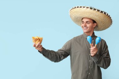 Marakas ve mavi arka planda bir kase nachos ile sombrero giyen yakışıklı genç adam.