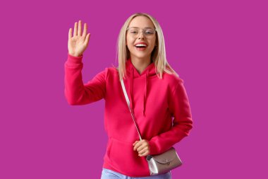 Şık kapüşonlu ve aksesuarlı güzel bir kadın mor arka planda el sallıyor.