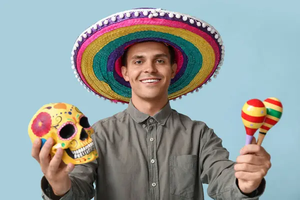 Handsome young man in sombrero with maracas and painted skull on blue background