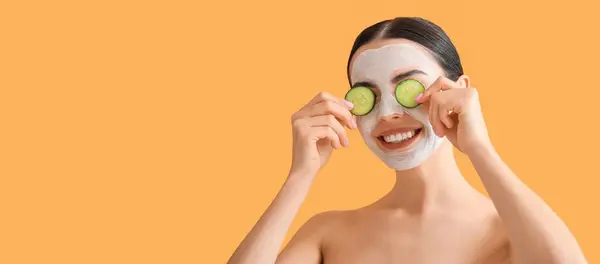 Beautiful young woman with cucumber slices and facial mask on orange background with space for text. Cosmetology concept