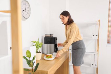 Güzel genç iş kadını ofiste lezzetli sandviçle kahve yapıyor.