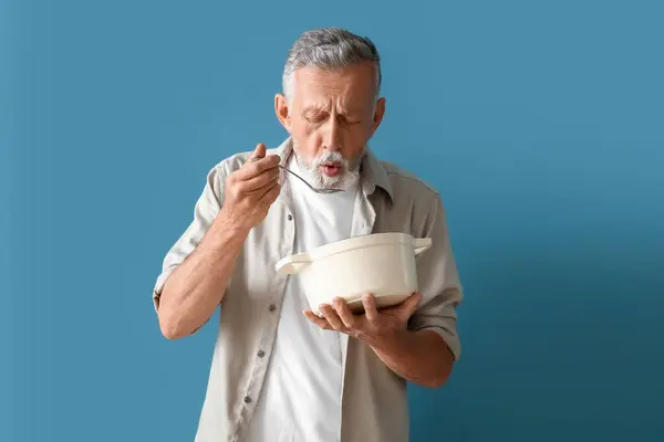 stock image Mature man with spoon and cooking pot on blue background